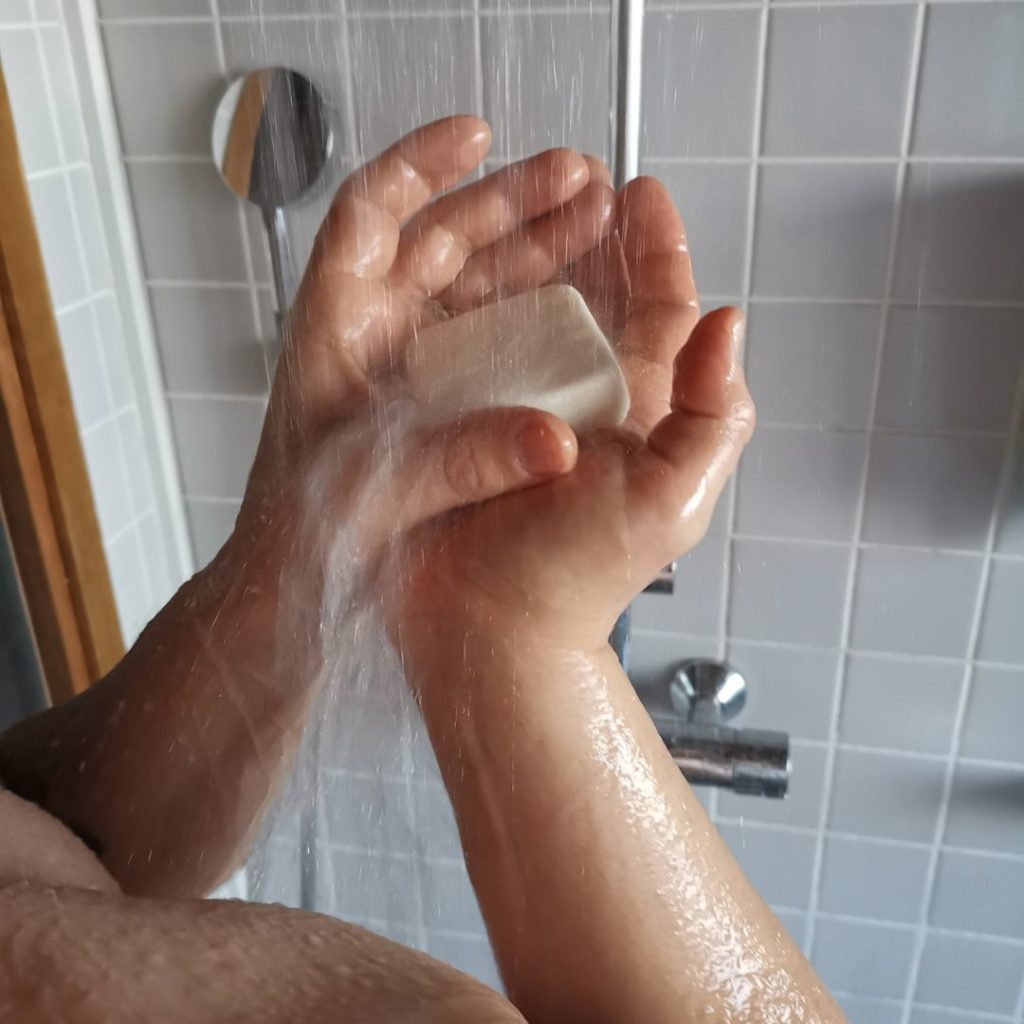 Kädet pitää palasaippuaa suihkun alla huuhtoutumassa